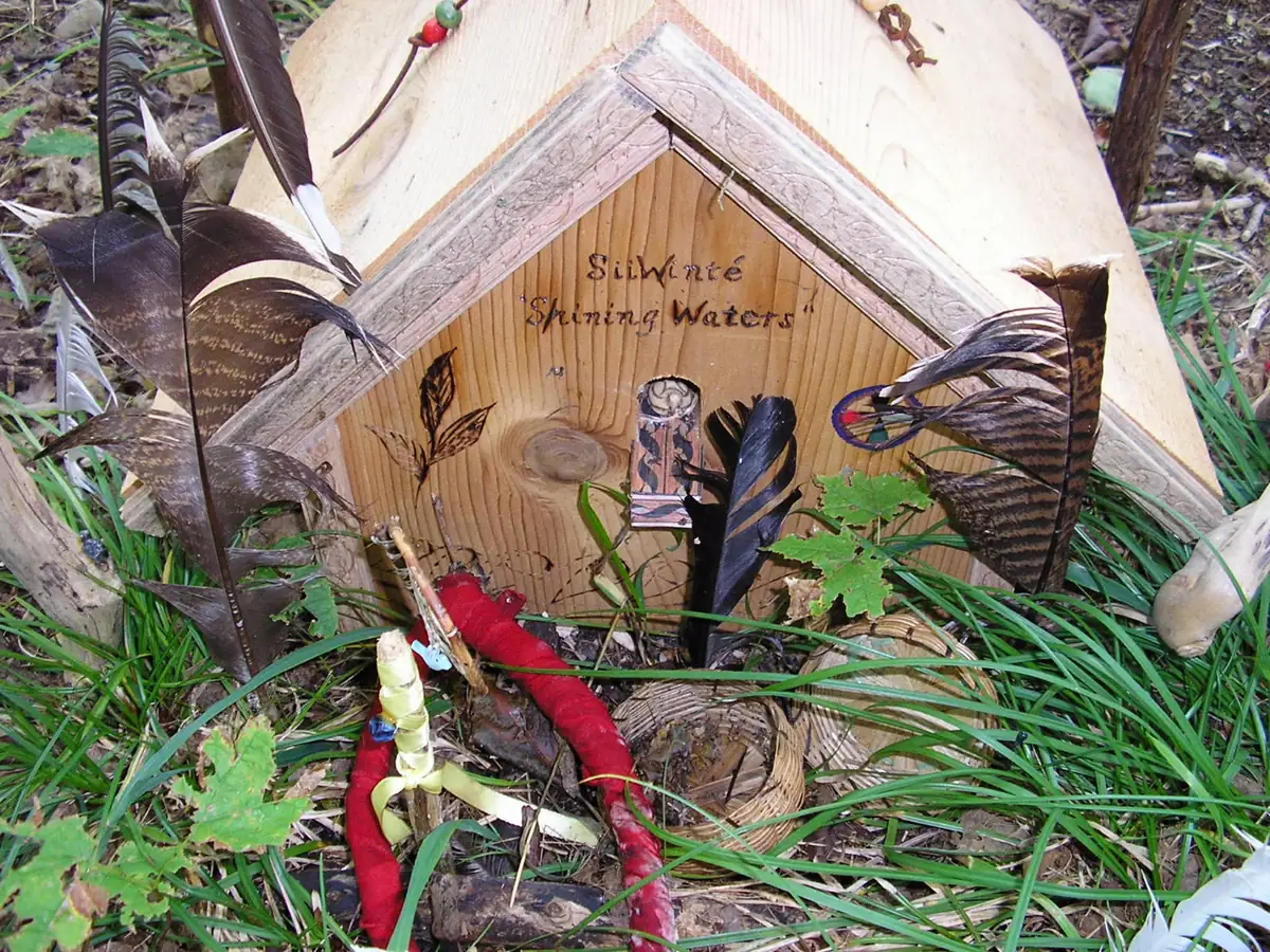 A decorated wooden structure with feathers, beads, red fabric, and the words “SiiWinté Shining Waters.”