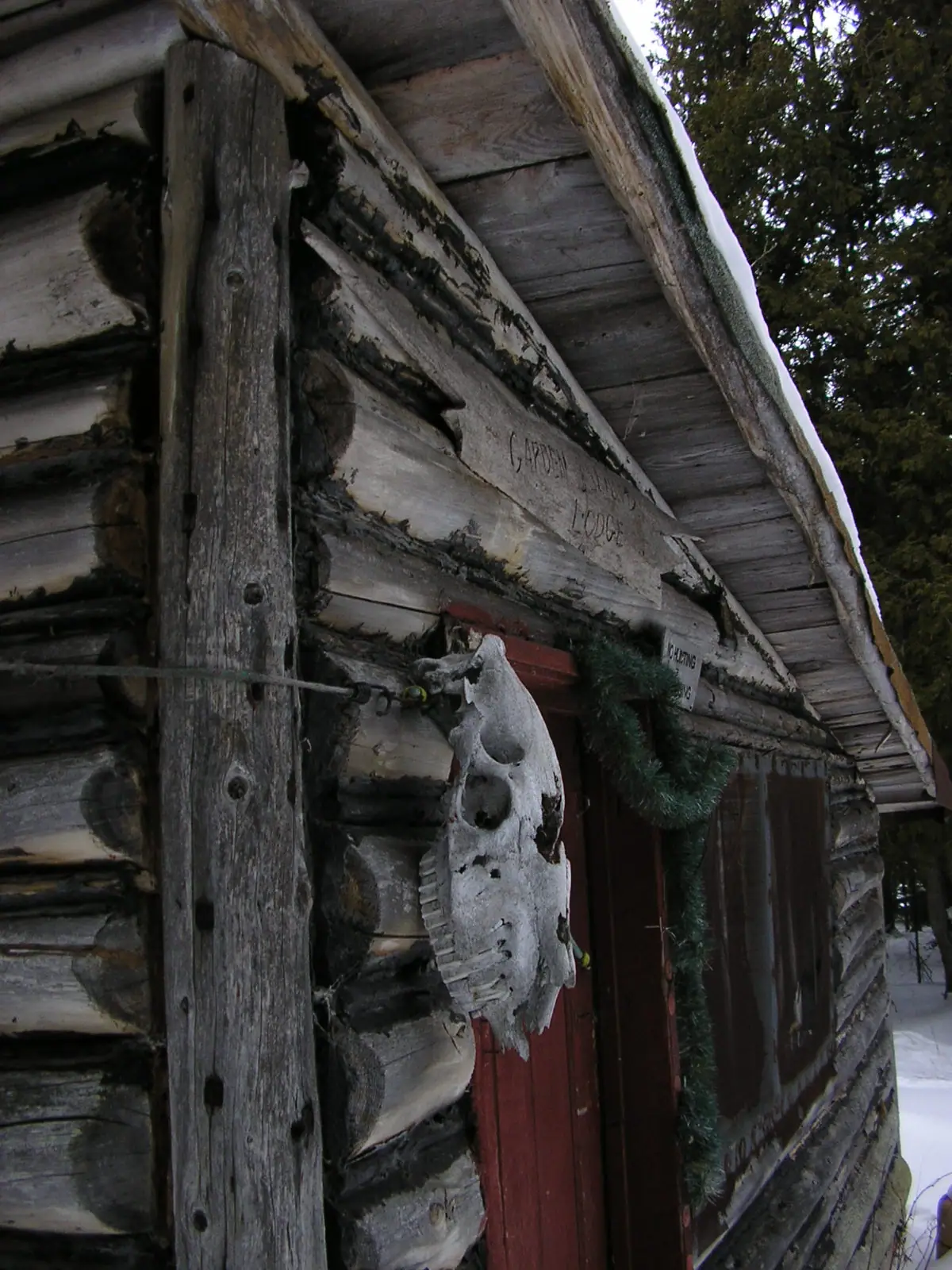 Snow-covered log building with a red door, evergreen garland, and animal skull mounted beside the entrance.