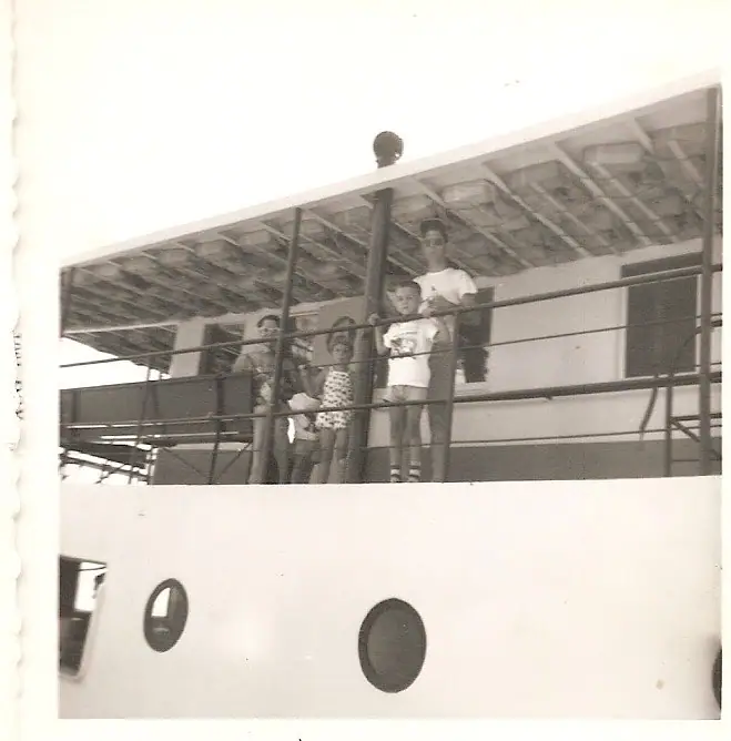 A group of adults and children stands behind a railing on the covered deck of a passenger vessel.