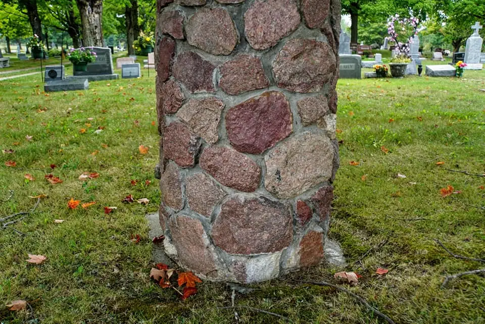 A stone-faced pillar stands among gravestones and flower arrangements in a grassy cemetery.