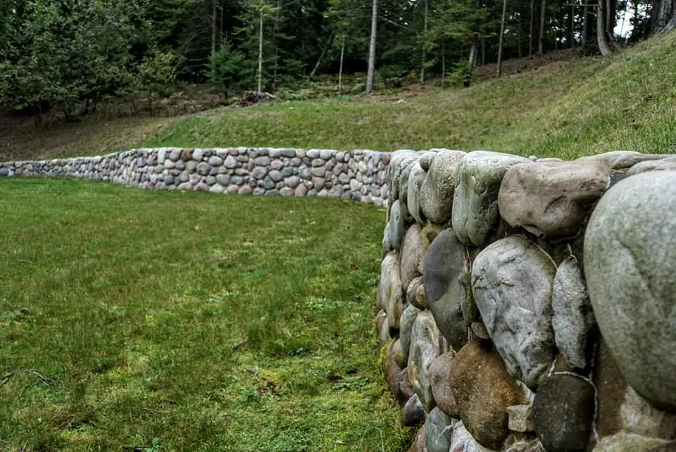 A rounded-stone wall curves across a grassy area beneath a wooded slope.