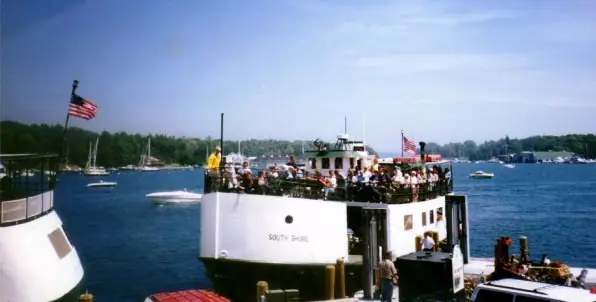 A crowded ferry named SOUTH SHORE approaches a ramp, with sailboats and wooded shoreline behind it.