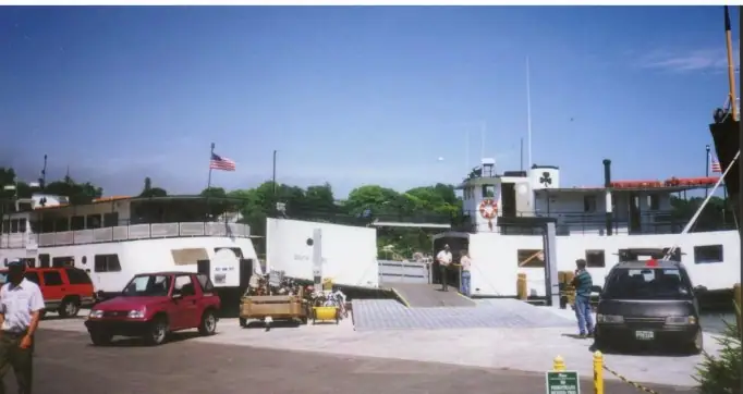 A white ferry is being loaded from a paved dock with vehicles, equipment, and people nearby.