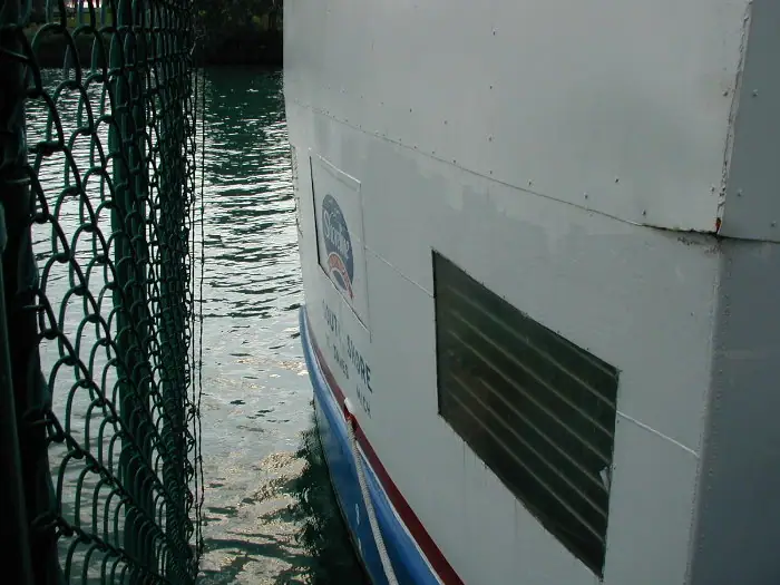 Close view of a white ferry stern beside a chain-link fence and rippling water.