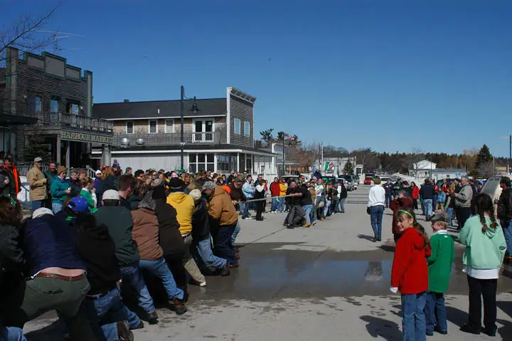 A crowd participates in a tug-of-war on a paved street beside storefronts and parked vehicles.