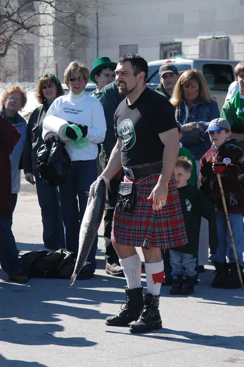 A kilted man holds a large fish while people and children watch outdoors.