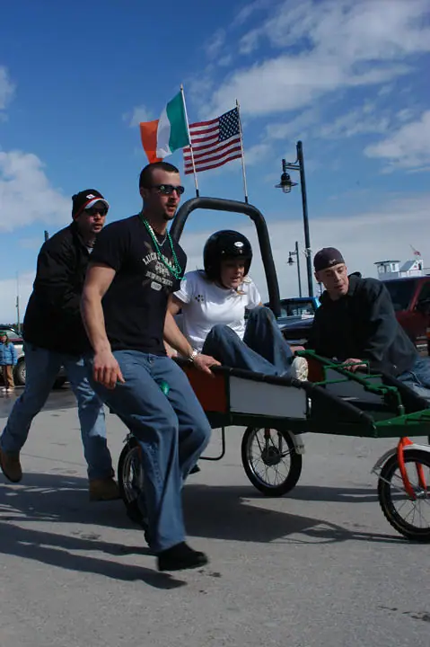 People ride and push a decorated pedal cart beneath Irish and American flags.