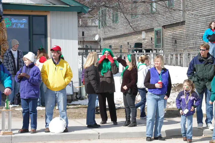 Adults and children gather outdoors beside a wooden building, with one person wearing a bright green wig.