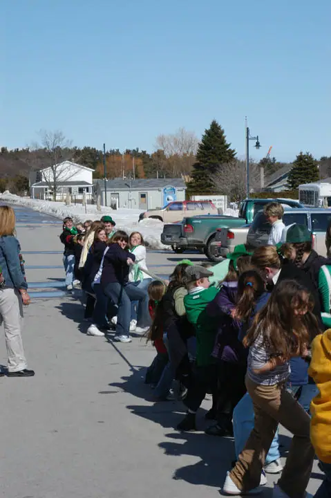 Children and adults pull a rope in a tug-of-war on a paved street beside snowbanks and parked pickup trucks.