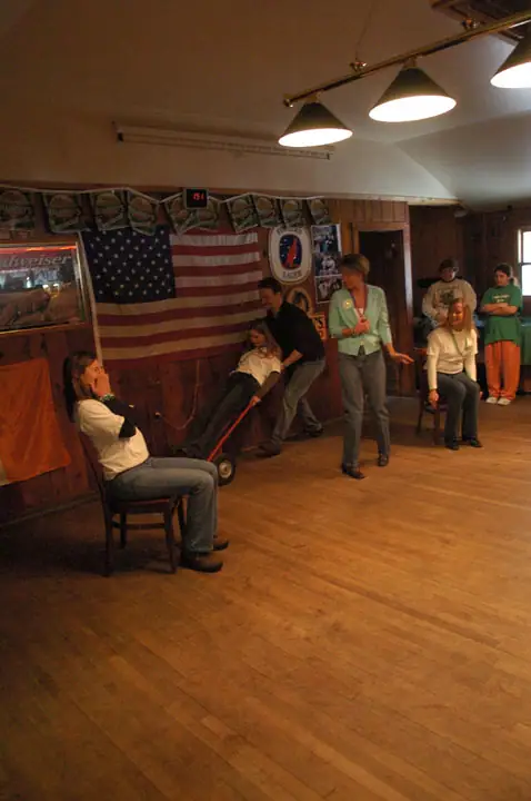 People play a party game in a wood-paneled room, with one person pushing another in a wheeled cart.