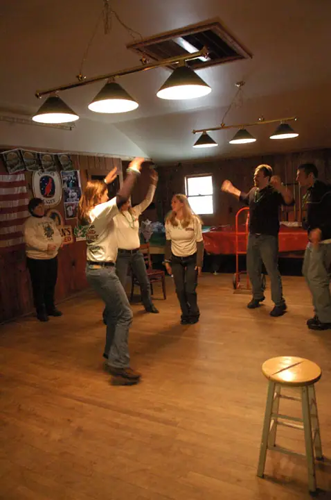 Six people dance in a wood-paneled room beneath hanging lights.