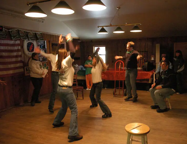 Adults dance in a wood-paneled room while others watch under bright ceiling lights.