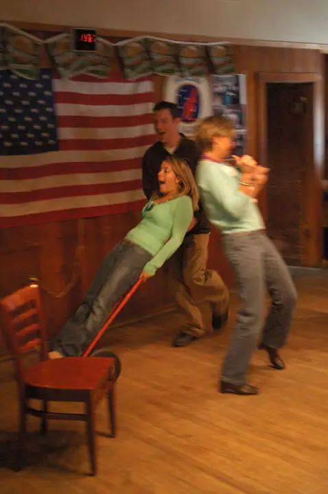 Three adults move playfully through a wood-paneled room decorated with an American flag.