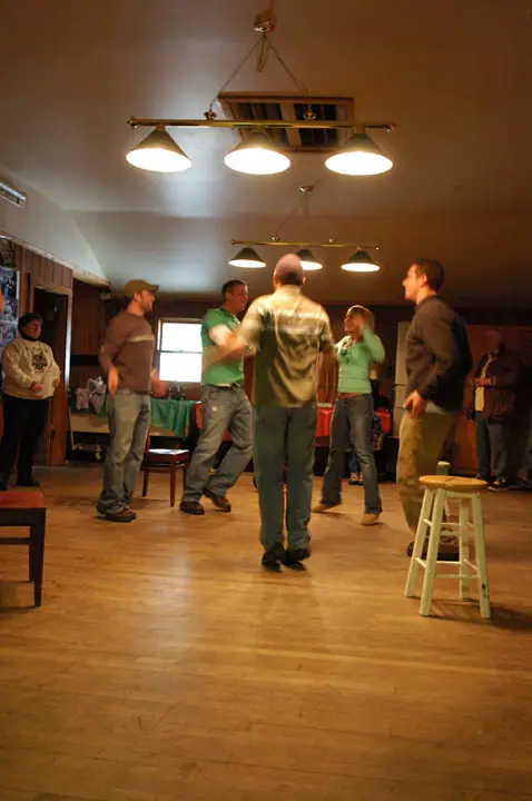 A group of adults dance in a wood-paneled room beneath hanging lights.