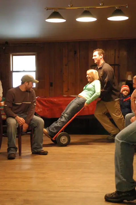 People playfully ride and push a red handcart inside a wood-paneled room during a gathering.