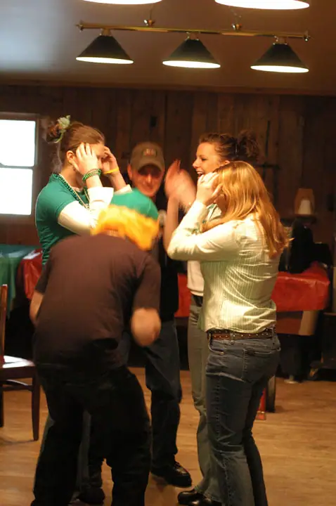 Several people gather indoors, with one person bent forward as others raise their hands around them.