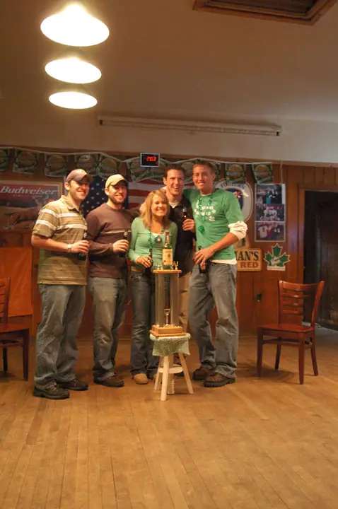 Five adults pose around a bottle on a small stand in a wood-paneled room decorated with beer signs and posters.