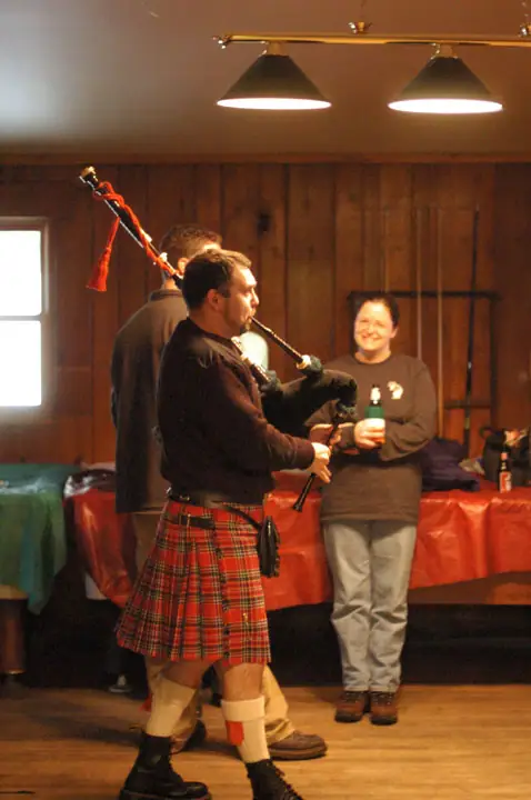 A kilted musician plays bagpipes while two people watch in a wood-paneled room.