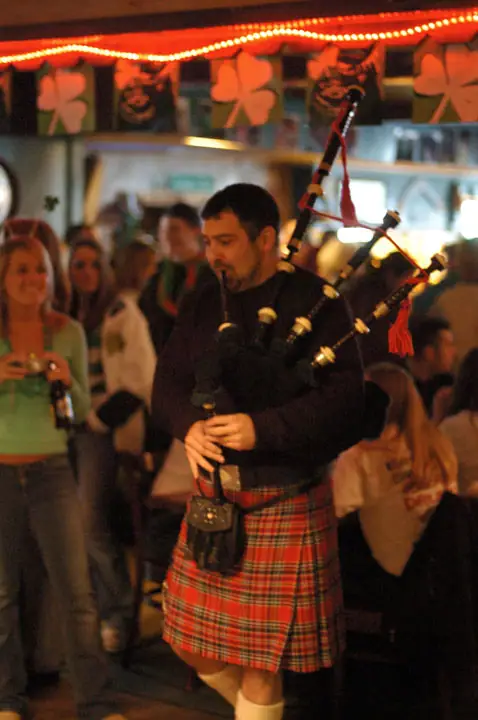 A bagpipe player in a red plaid kilt performs for a crowd in a warmly lit decorated interior.