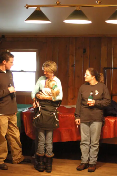 Four people gather indoors, with an adult holding a child beside a red-covered table.