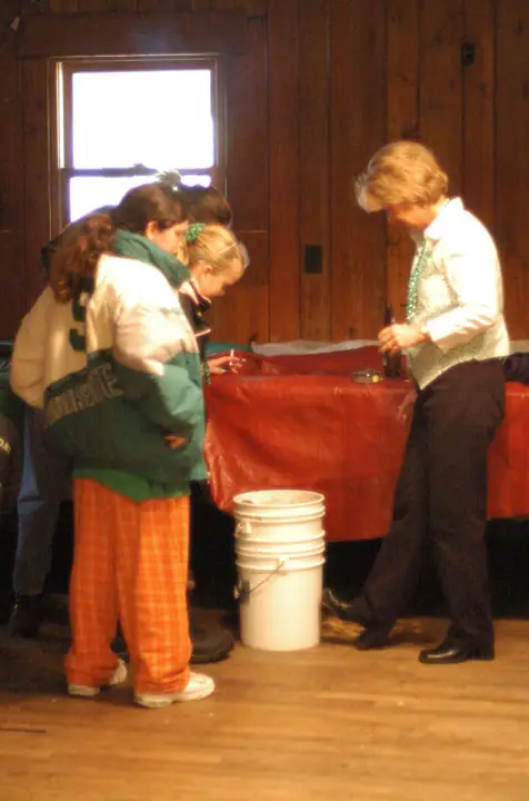 Four people gather around a red-covered table in a wood-paneled room, with a white bucket on the floor.