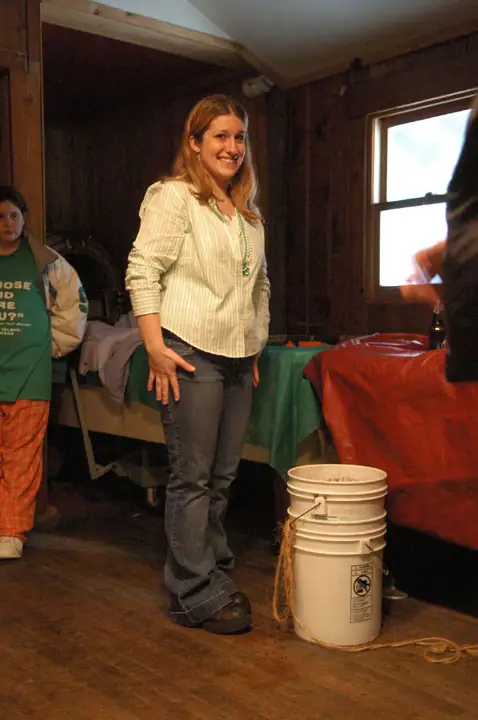 A smiling woman stands indoors beside another person, tables, stacked buckets, and a window.