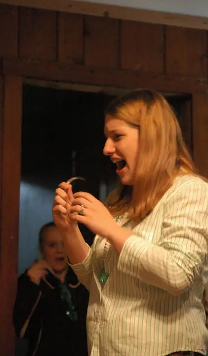 A woman holds a small curved object while another person watches from a dark wooden doorway.