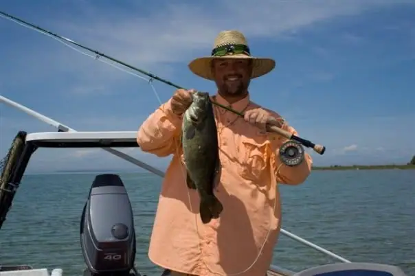 Smiling angler in a straw hat holds a large fish and fishing rod aboard a motorboat.