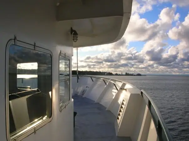 View from a white vessel’s passenger deck toward a low shoreline beneath a partly cloudy sky.