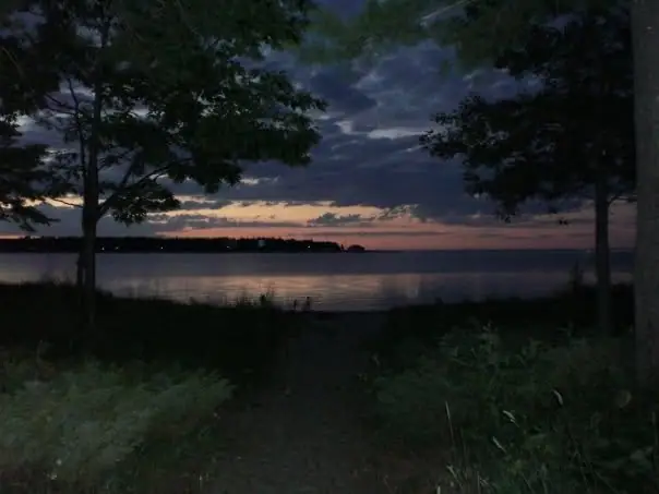 A dusky sky glows over calm water, framed by trees and a narrow path through shoreline vegetation.