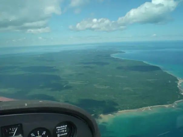 Aerial view of a forested coastal landform and bright blue water seen from an aircraft cockpit.