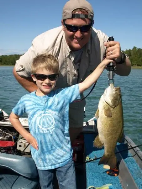 An adult and child pose on a boat while holding a large fish suspended from a hand grip.