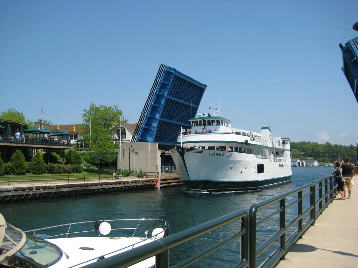 A white passenger ferry passes beneath a raised blue drawbridge beside a waterfront walkway.