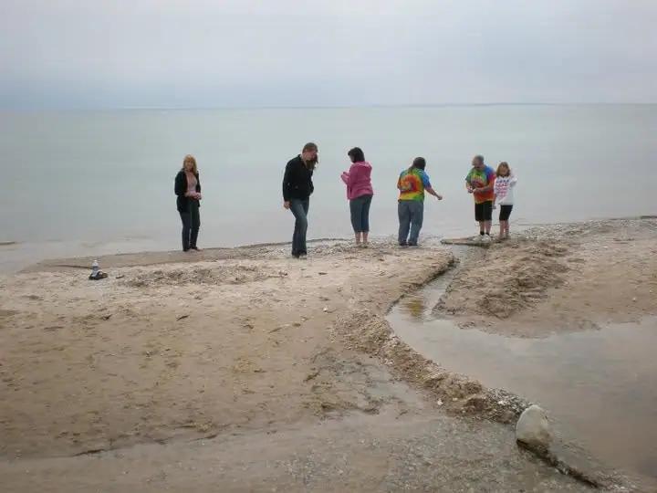 Six people stand on a sandy shoreline beside shallow flowing water under an overcast sky.