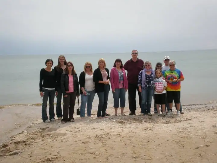 A group of adults and children poses on a sandy, pebbly shoreline with calm water behind them.