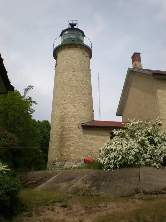 A cylindrical brick lighthouse and adjoining red-roofed building stand beside flowering shrubs and trees.