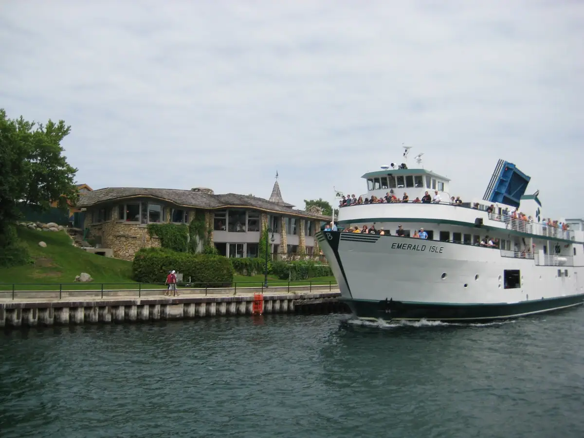 A white ferry carrying passengers passes a dock beside a low stone building with a shingled roof.