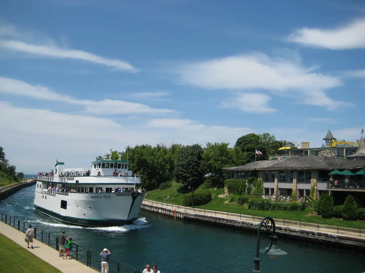A crowded white passenger ferry moves through a narrow channel beside a walkway and stone-faced waterfront business.