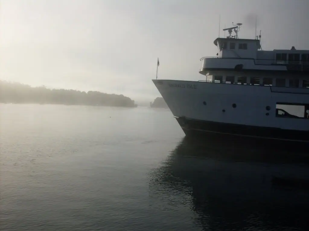A large white ferry emerges through mist on calm water beside a wooded shoreline.