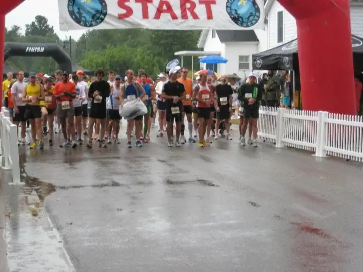 Runners gather beneath a START banner on a wet street beside a white building and picket fence.