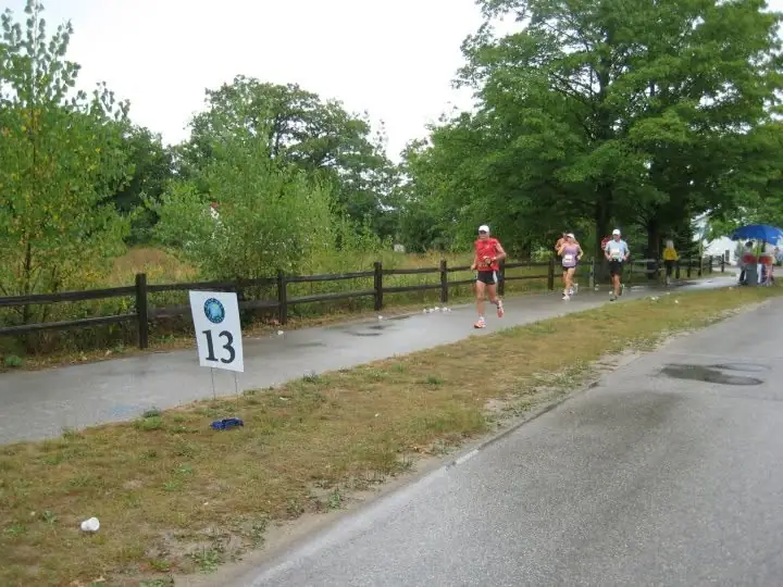 Runners pass a roadside marker numbered 13 on a wet path beside a wooden fence.