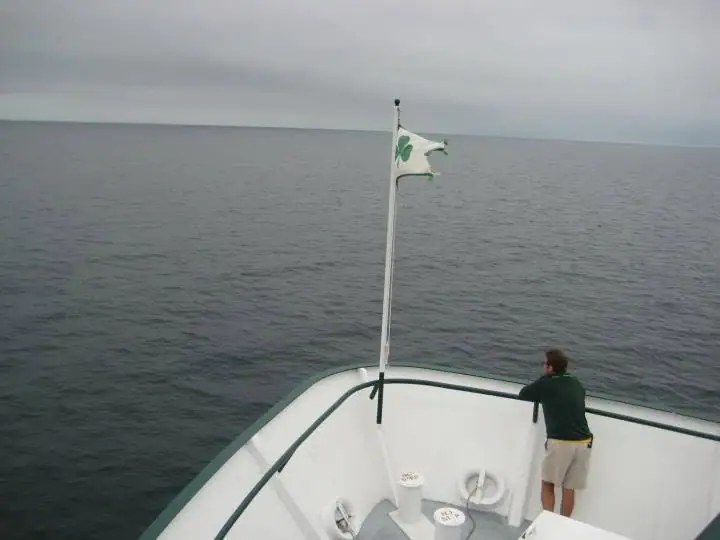 A person stands at the bow of a passenger vessel beneath a white shamrock flag on dark water.