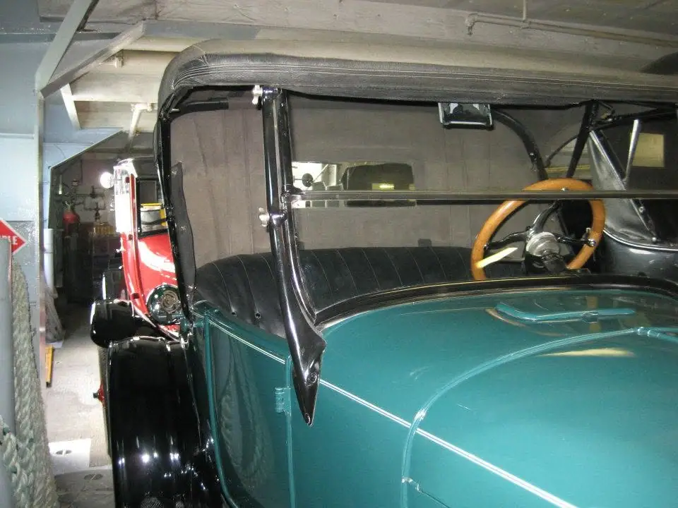 A teal vintage automobile with black fenders and upholstery is parked inside a garage beside a red vehicle.
