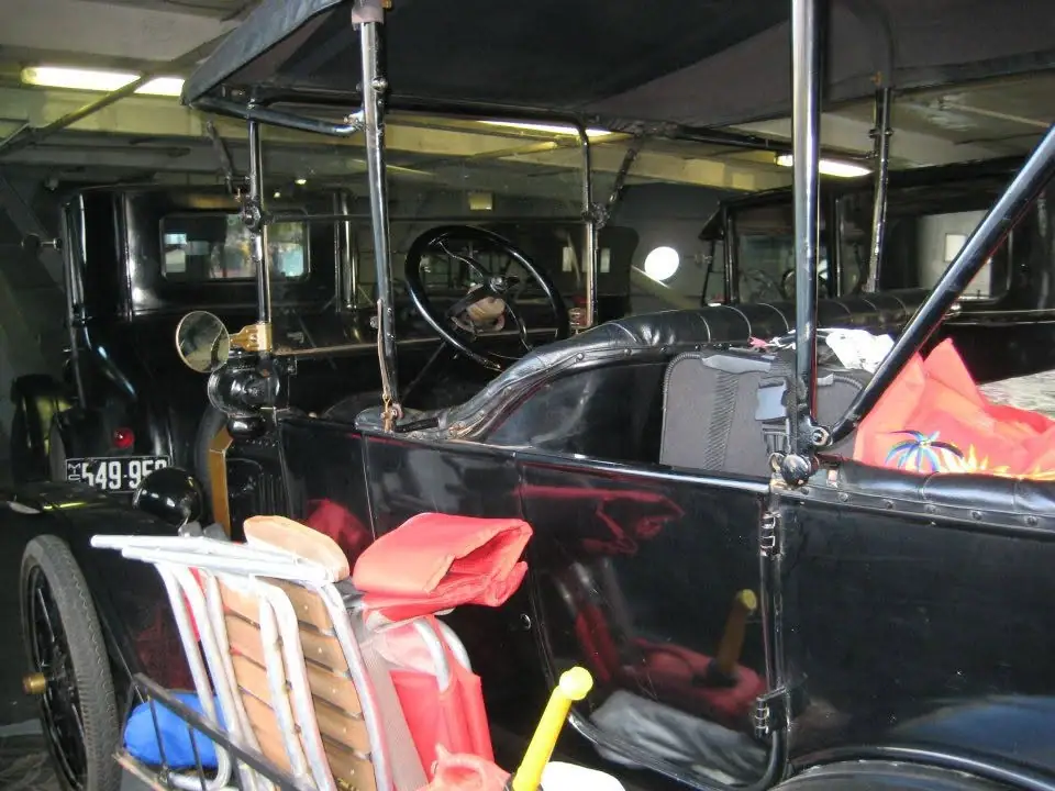 A black antique open-top automobile is parked indoors beside another vintage vehicle and folding chairs.