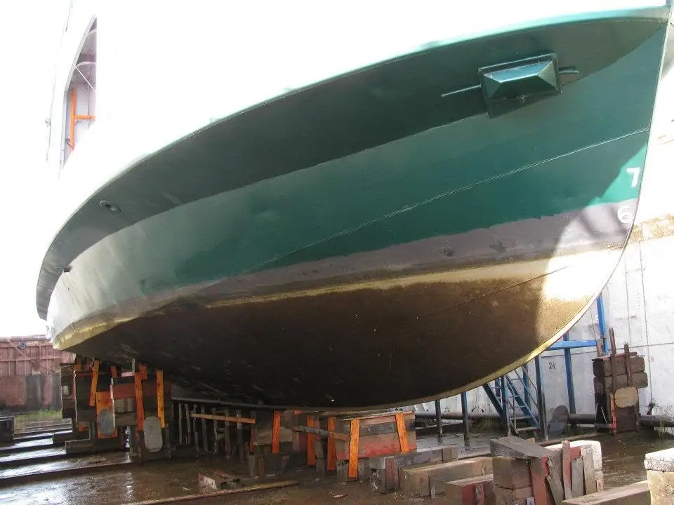 A large green vessel hull rests on wooden supports in a wet shipyard or dry dock.