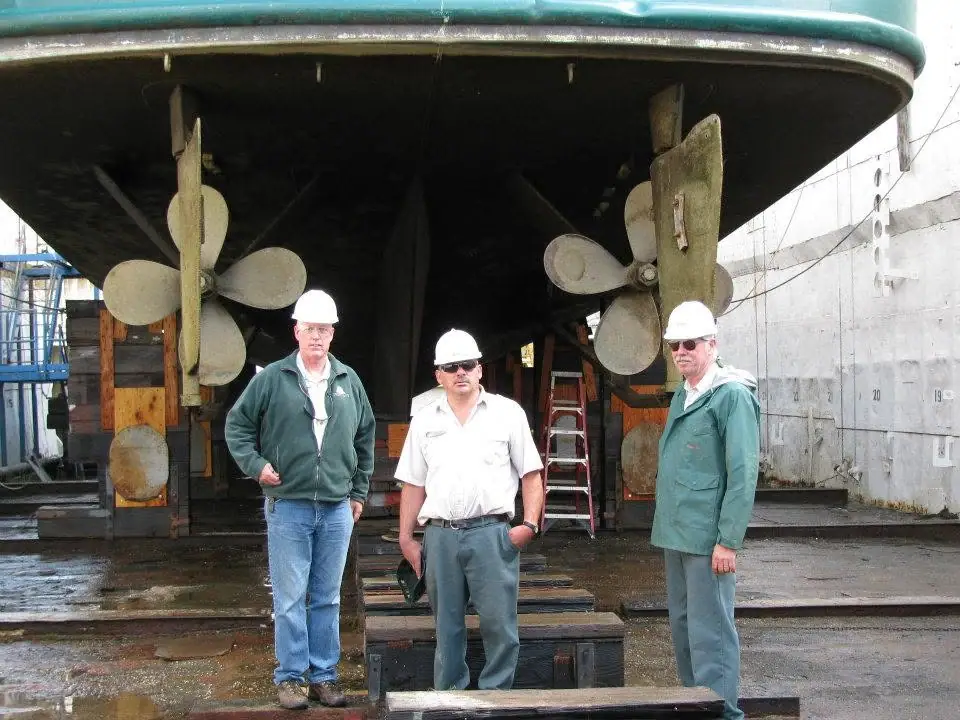 Three men in hard hats stand beneath a large vessel’s stern and twin propellers.