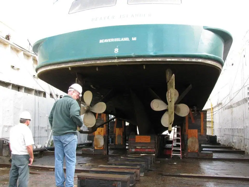 The stern of a green ferry raised in a dry dock, with two workers beside its propellers.
