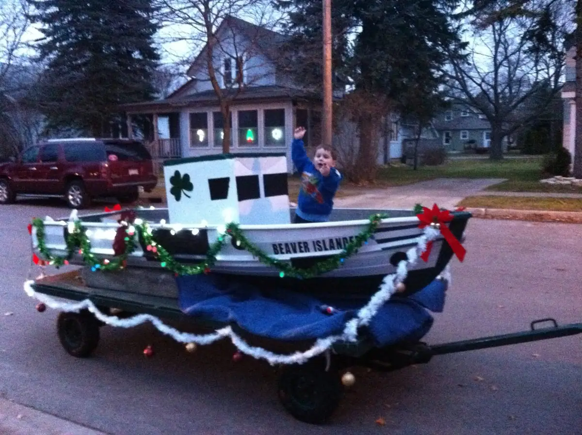 A child waves from a small boat-shaped parade float decorated with garlands, lights, and red bows.