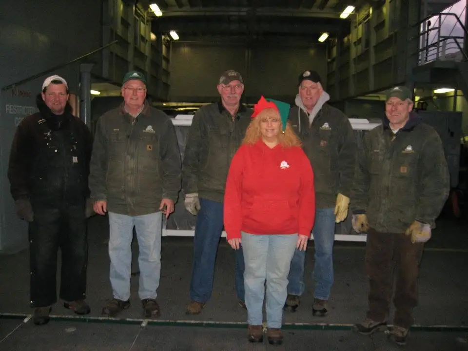 Six workers pose indoors, with one wearing a red holiday hat and several wearing work jackets and gloves.