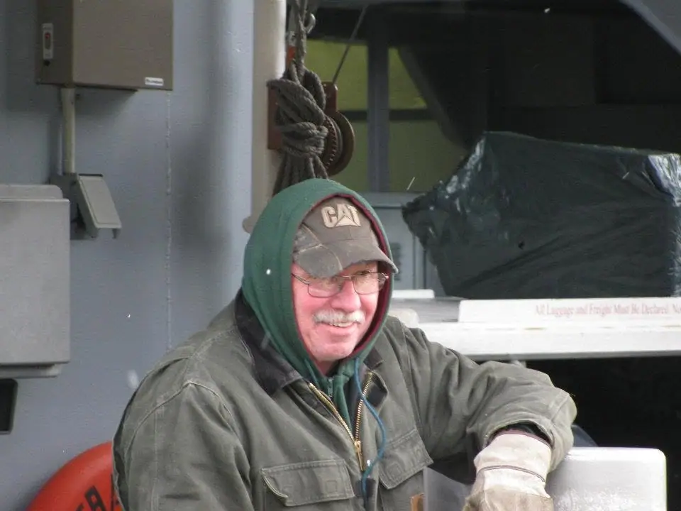 Smiling man in a hooded jacket and CAT cap seated beside cargo equipment aboard a vessel.
